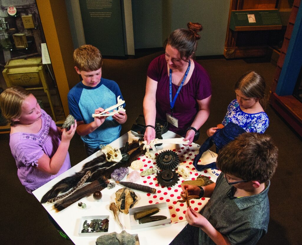 A woman and children gather around a table filled with various items, looking curiously at what's displayed.  