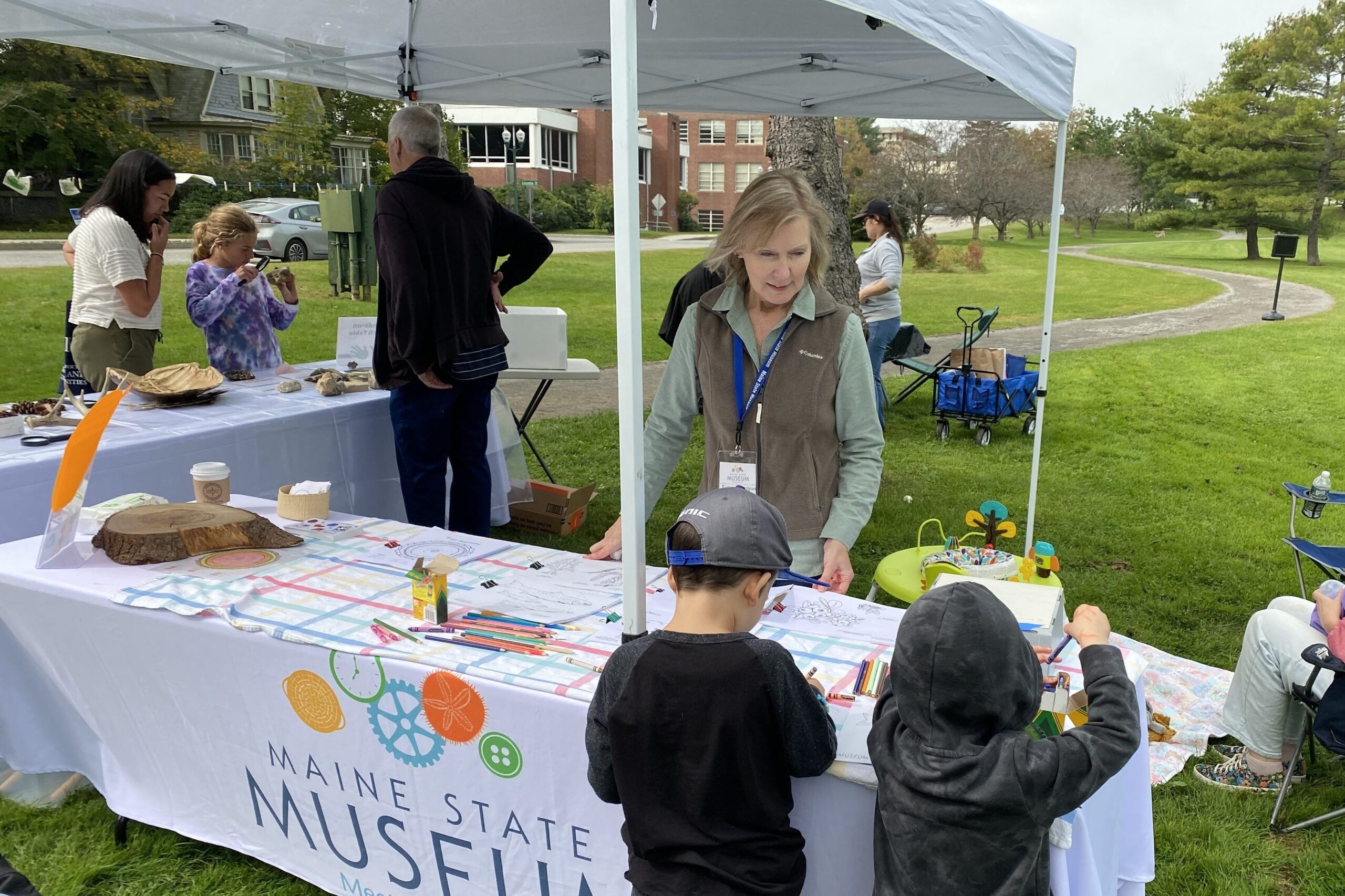 Smiling woman helps children with an art project at an outdoor table.